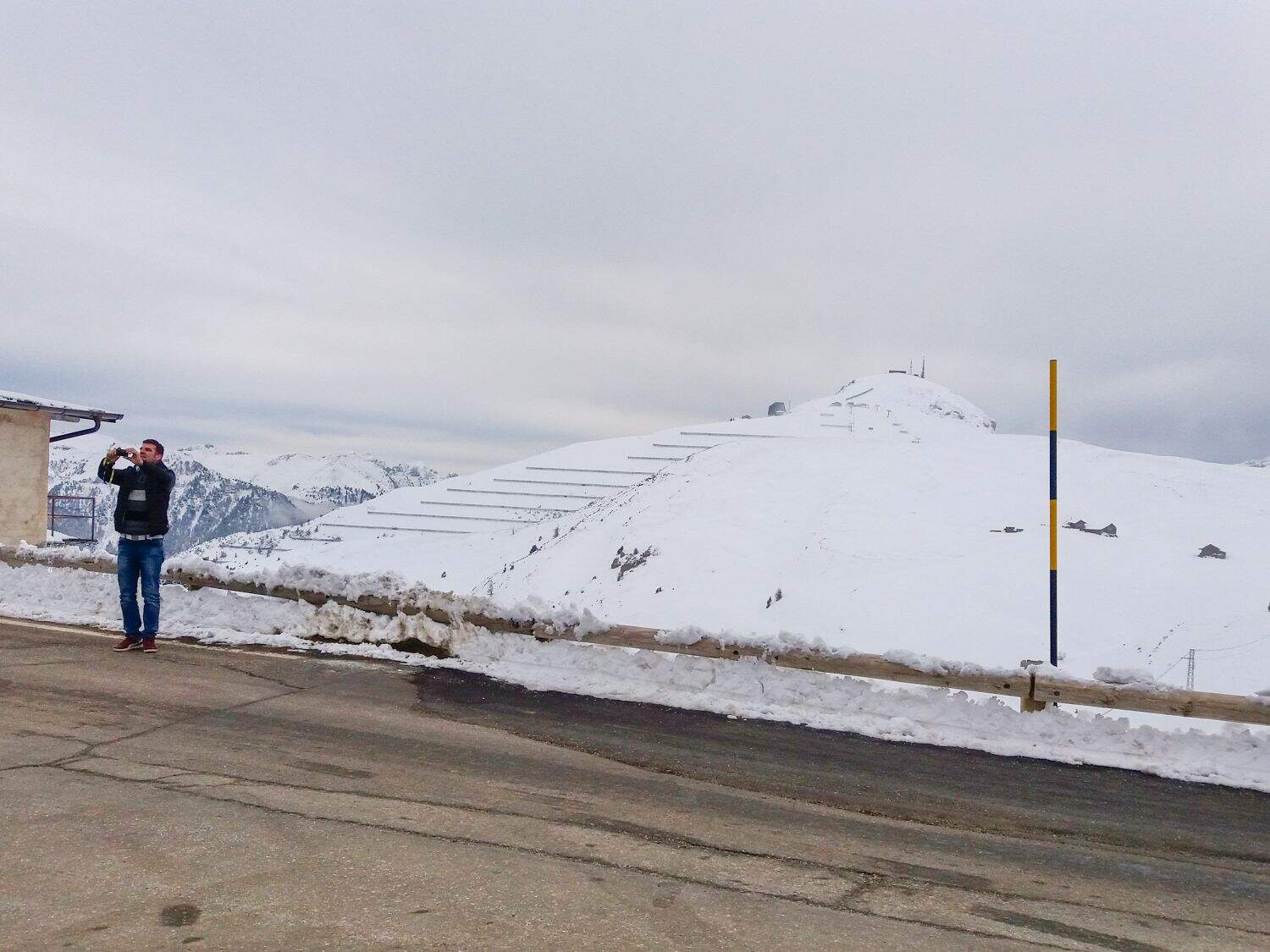 A person stands on the side of a paved road, taking a photo of the snowy mountain landscape. Snow covers the ground, and the sky is overcast. Steps are visible on the slope in the background.