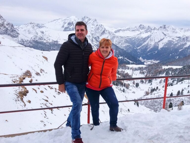 A man and woman stand smiling together on a snowy mountain viewpoint with snow-covered peaks and trees in the background. Both wear jackets, the woman in orange and the man in black, enjoying the winter scenery.
