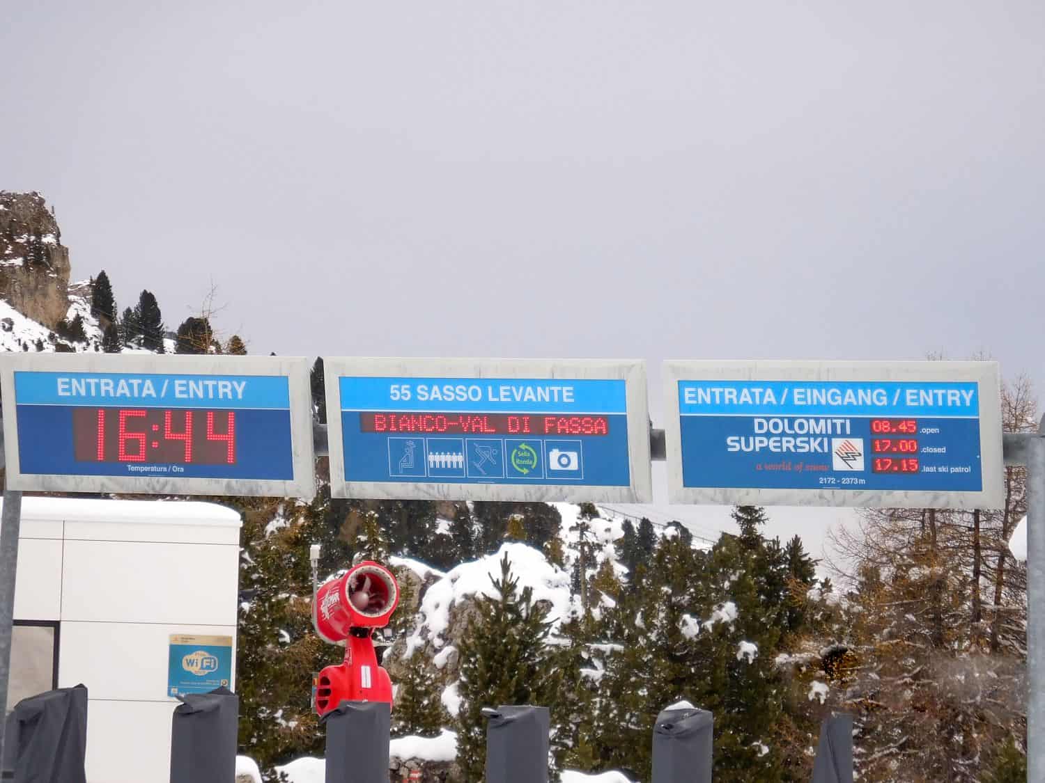 Electronic ski resort signs display entry times, ski lift information, and Dolomiti Superski prices in Italian, German, and English, with trees and snow-covered mountains in the background.