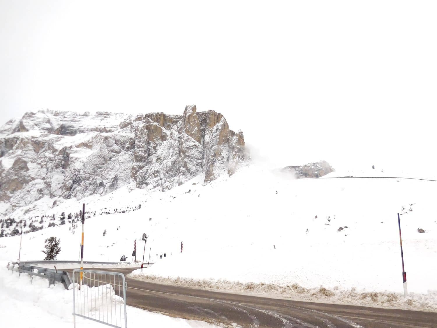 A snow-covered mountain rises behind a winding road, partially lined with a metal barrier. The scene is overcast with white sky, blending with the snowy ground, creating a wintry and serene landscape.