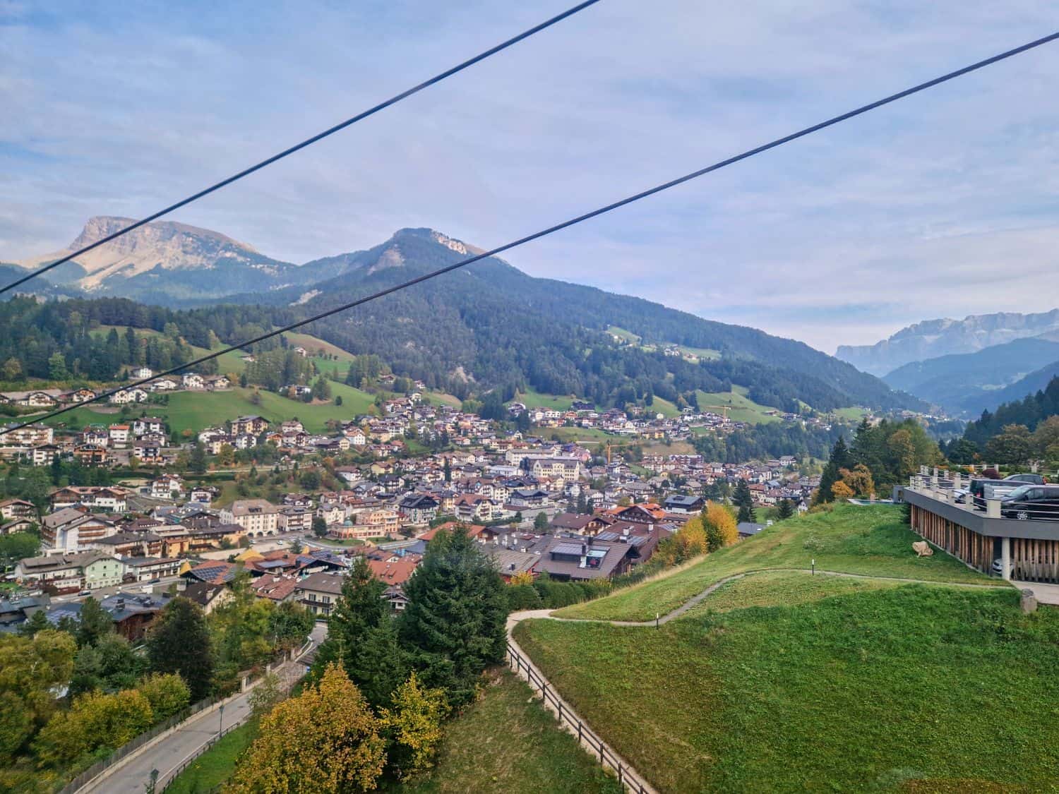 View of a small mountain town with clustered houses, green hills, and forested mountains. Two cables run across the sky, and a modern building sits on a grassy slope in the foreground.