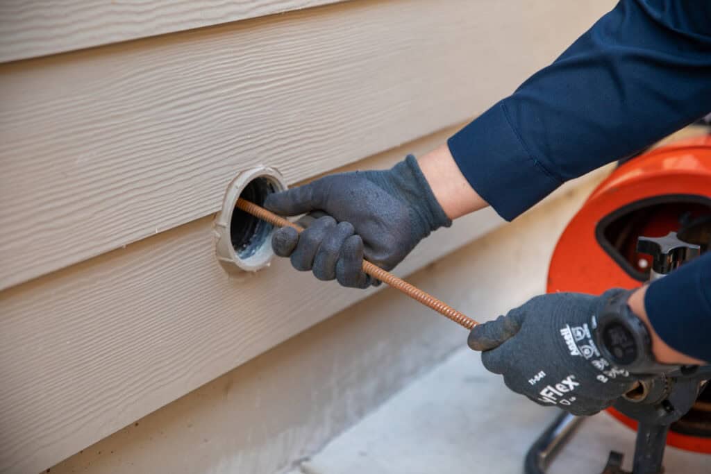 A Moore Home Services pro in gloves expertly clears a home’s exterior drain pipe with a snake—guaranteeing fast, tidy service.