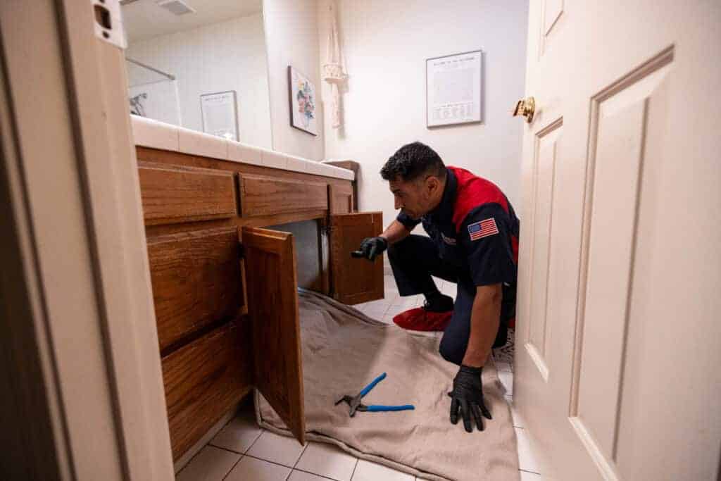 A friendly, uniformed plumber from Moore Home Services kneels on a spotless bathroom floor, expertly inspecting pipes beneath the sink. Wearing protective gloves and keeping tools neatly organized on a towel, he demonstrates our commitment to thorough fall plumbing maintenance before the cold arrives. Trust us for reliable, guaranteed plumbing and drain service—always leaving your home as clean as we found it.