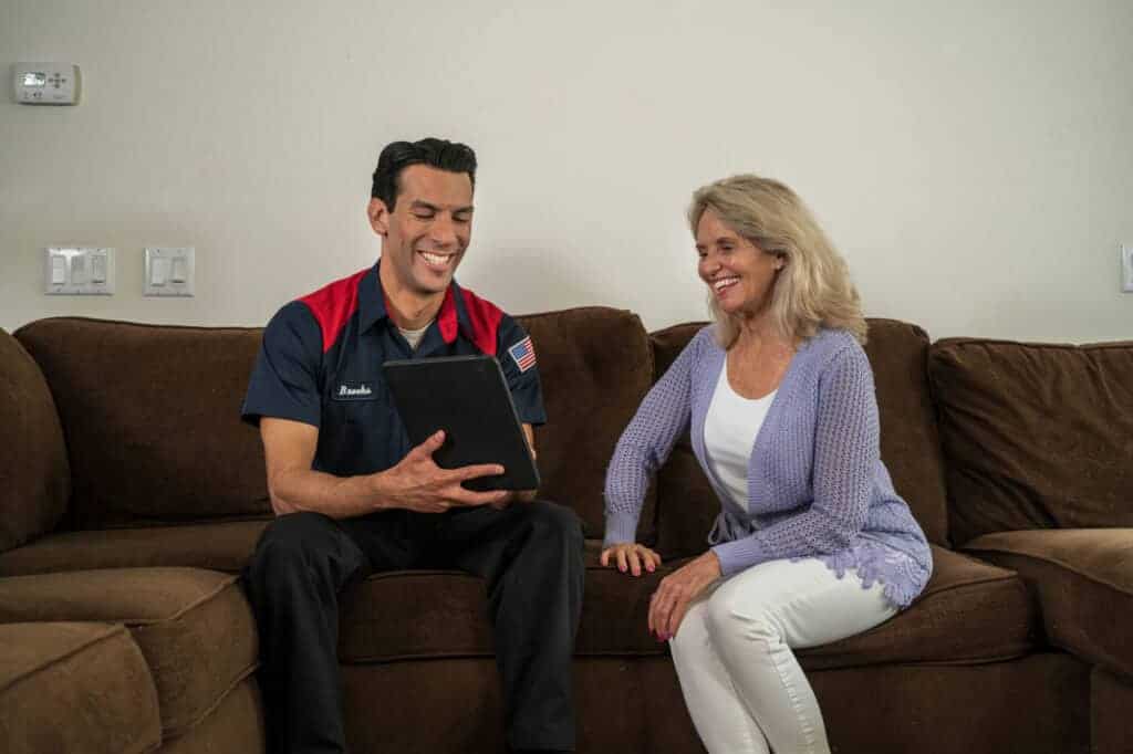 A friendly technician in a Moore Home Services uniform sits with a smiling homeowner on a cozy brown couch, using a tablet to share expert advice on programmable thermostats and how they can boost home comfort and energy savings. Our trusted HVAC professionals are here to deliver clean, reliable service backed by our satisfaction guarantees—because your comfort is always our top priority.
