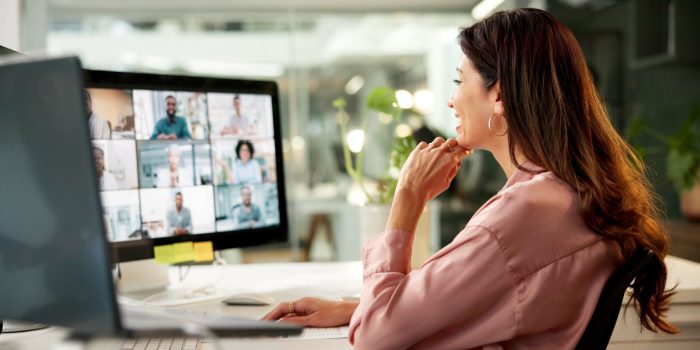 A businesswoman at her desk views a grid of virtual meeting participants on her desktop monitor, illustrating the modern remote work setting where AI notetaking tools are commonly deployed.