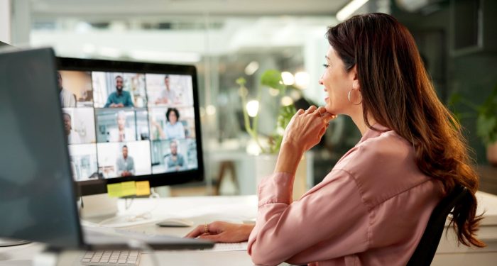 A businesswoman at her desk views a grid of virtual meeting participants on her desktop monitor, illustrating the modern remote work setting where AI notetaking tools are commonly deployed.