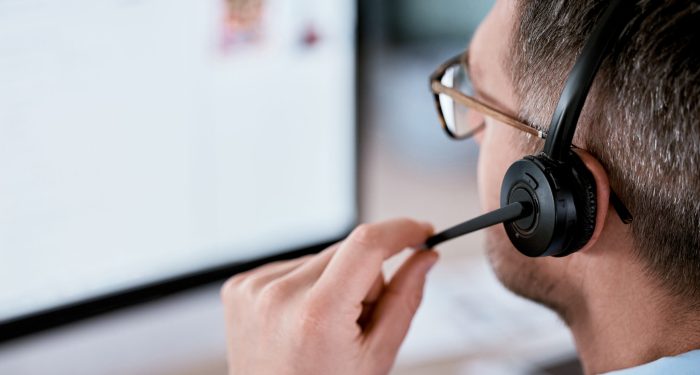 Call center agent wearing a headset and glasses adjusts his microphone while looking at a computer screen.