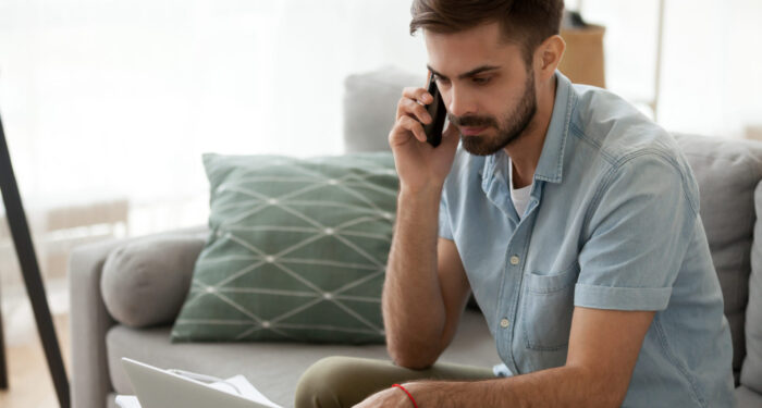 Man sitting on couch reading message email online looking at laptop talking by mobile phone. Male working at home has serious conversation with client. Businessman solve problems and matters by phone