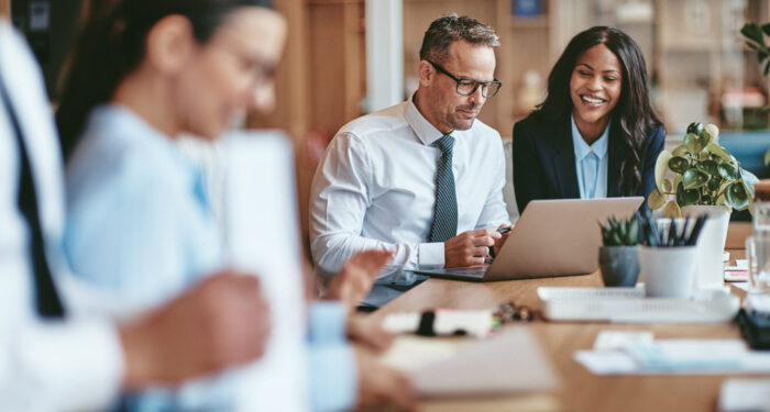 Two diverse businesspeople smiling while working on a laptop together at the end of a boardroom table in an office