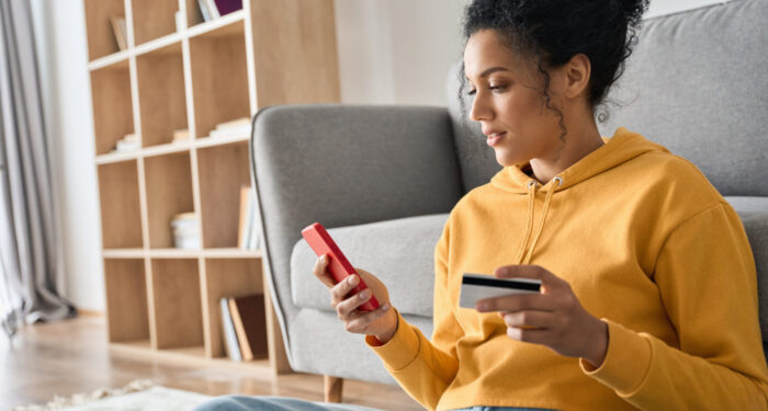 Young adult African American female consumer holding credit card and smartphone sitting on floor at home doing online banking transaction. E commerce virtual shopping, secure mobile banking concept.
