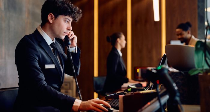 Young receptionist making a phone call while working on desktop PC at hotel reception desk.
