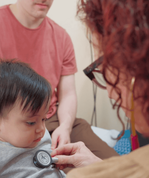 A baby in a grey shirt looking down at his chest while a doctor listens to his heart rate with a stethoscope in a patient clinic setting and parents in the background.