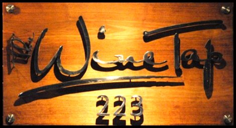 A metal sign on a wood wall is lit from above, creating deep shadows of the letters that read, “Wine Tap 223.”
