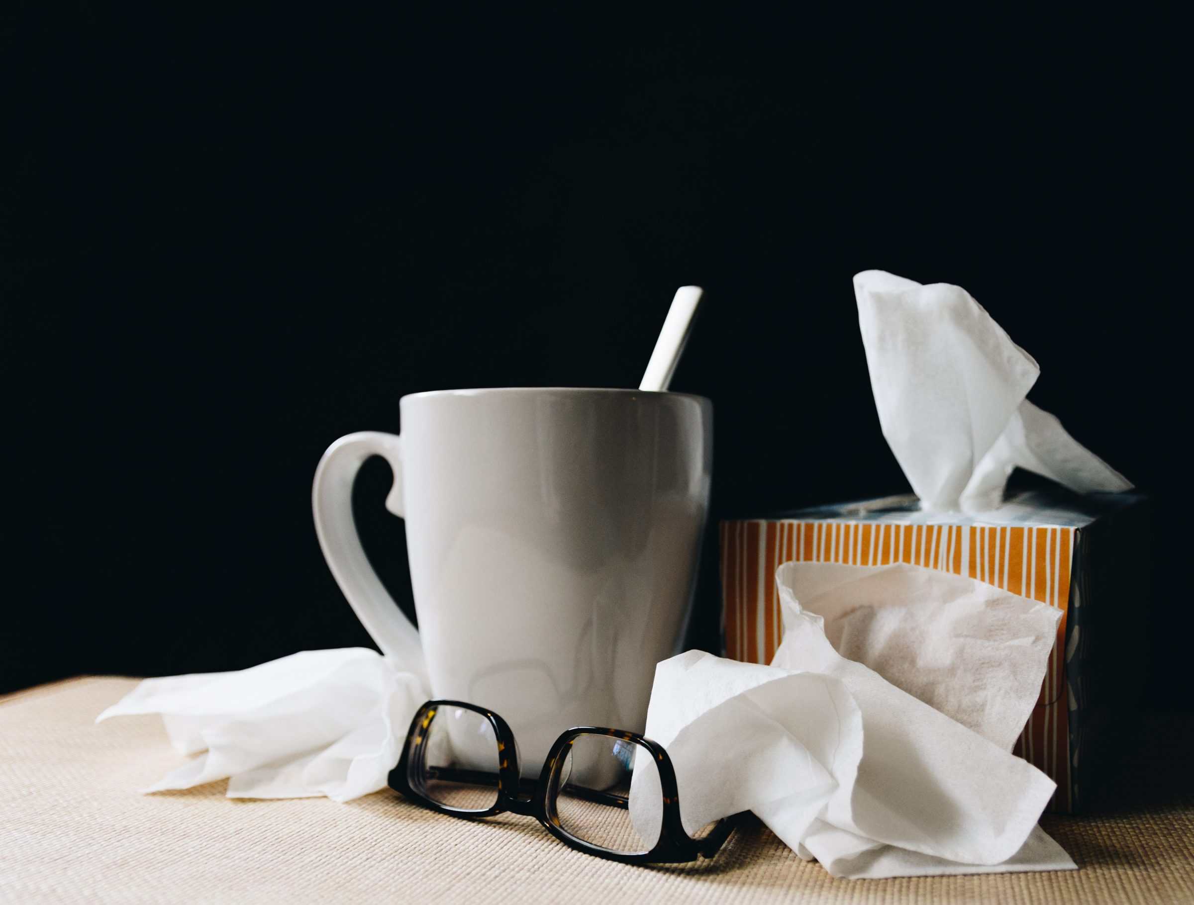 A spoon in a white mug, a box of tissues, and reading glasses sit on a table against a black backdrop illustrating how dry air in winter can increase sickness.