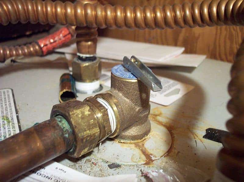 A photo of a gold-colored water-heater valve. There is a copper coil behind the valve, and the valve connects to the top of a white water heater tank. 