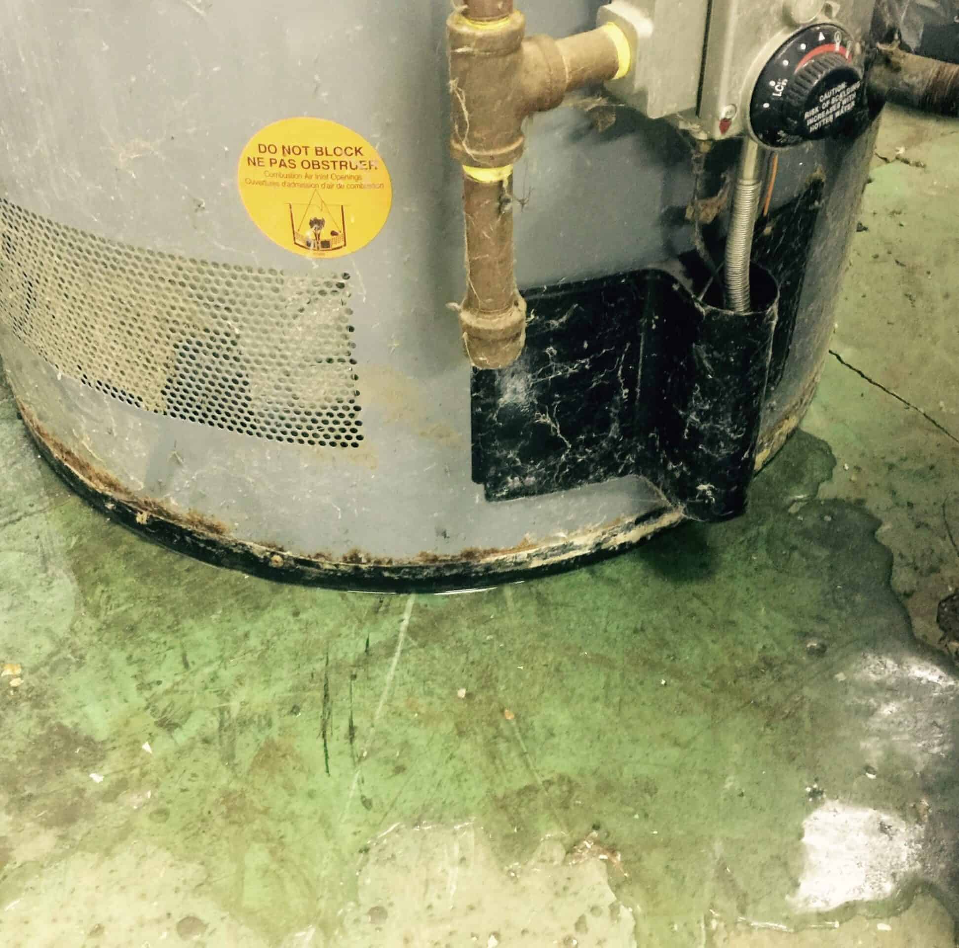 A green tile floor is covered in water beneath a leaking hot water heater. The water heater is grey colored with gold and black pipes.