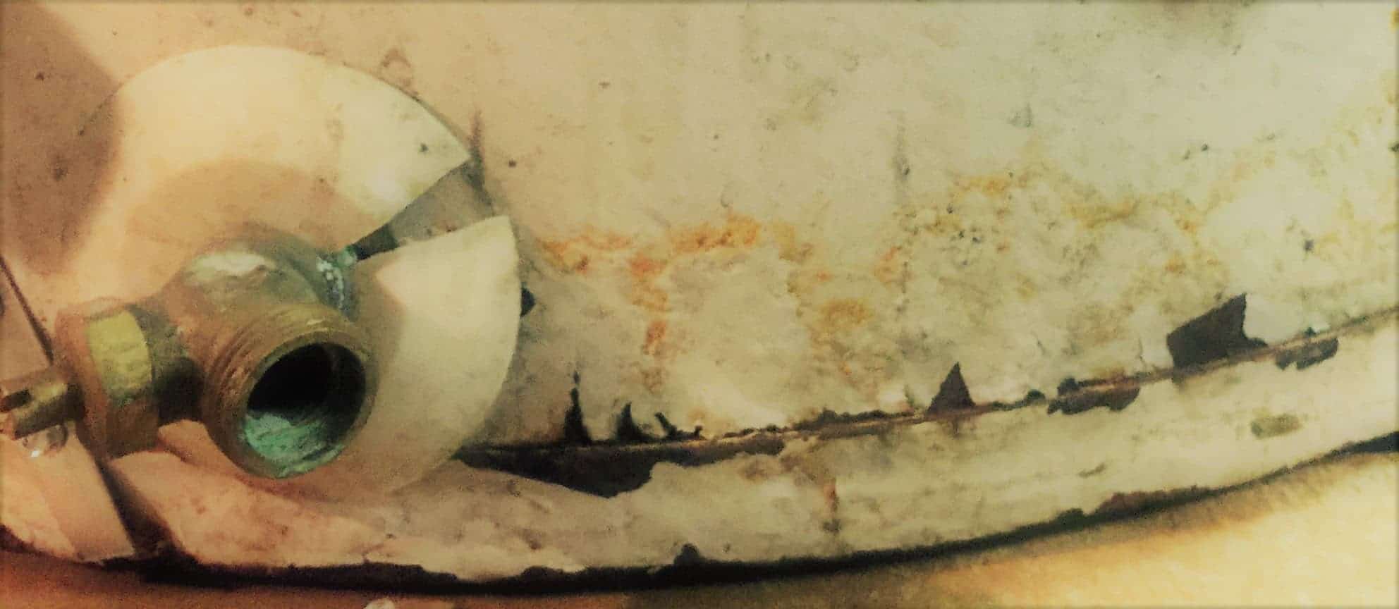 A white colored water heater displays a rusty crack along the bottom. A brass fitting shows rust and tarnish from a water leak.
