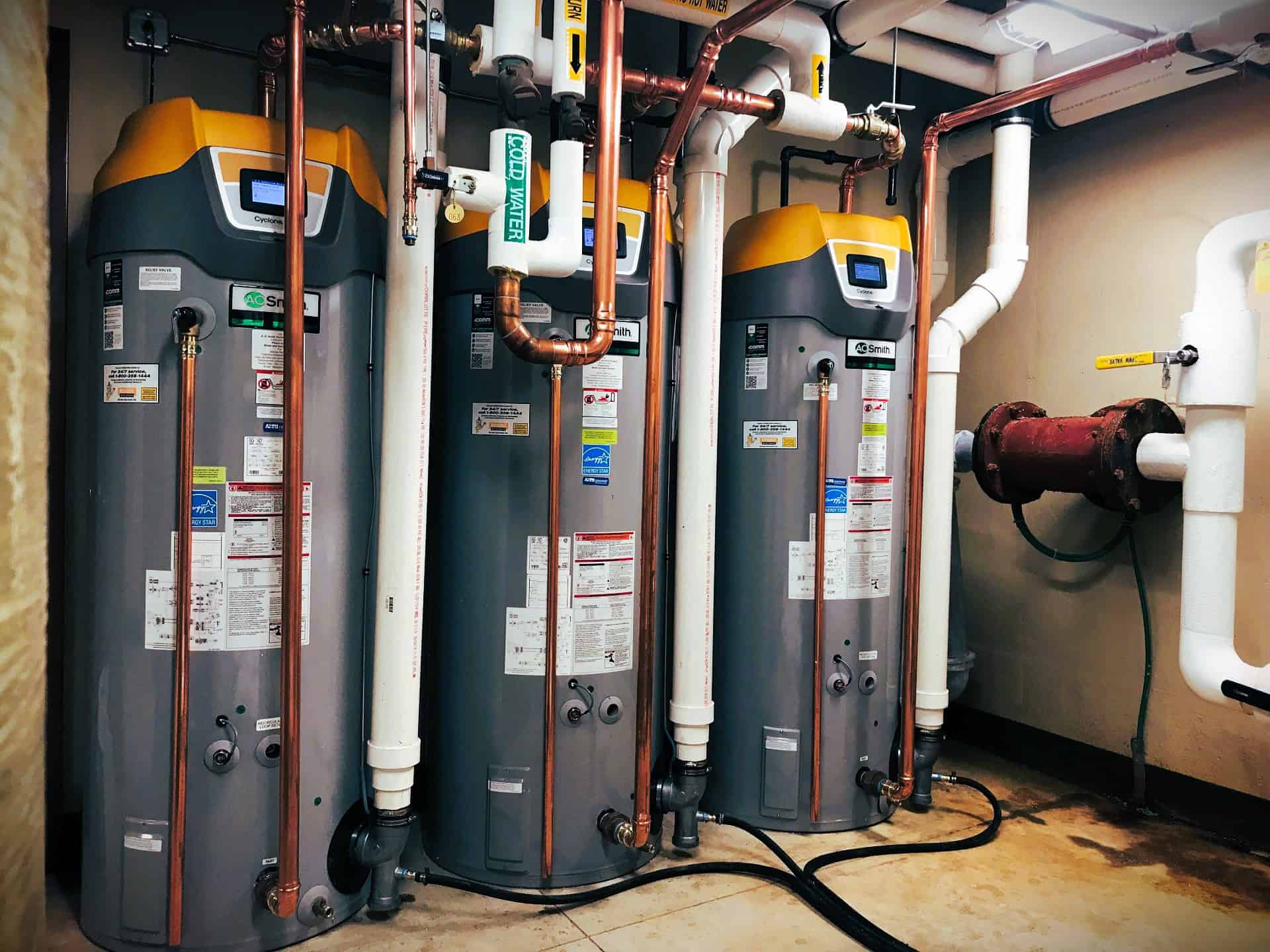 Three large water heaters setup for redundancy. Each water heater is connected to large white PVC pipes and thinner copper pipes.