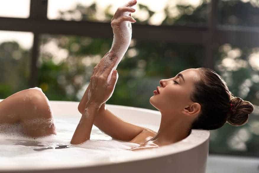 A woman relaxes in the bathtub next to large windows, her hands soapy and free of dryness, thanks to the many benefits of a water softener. 