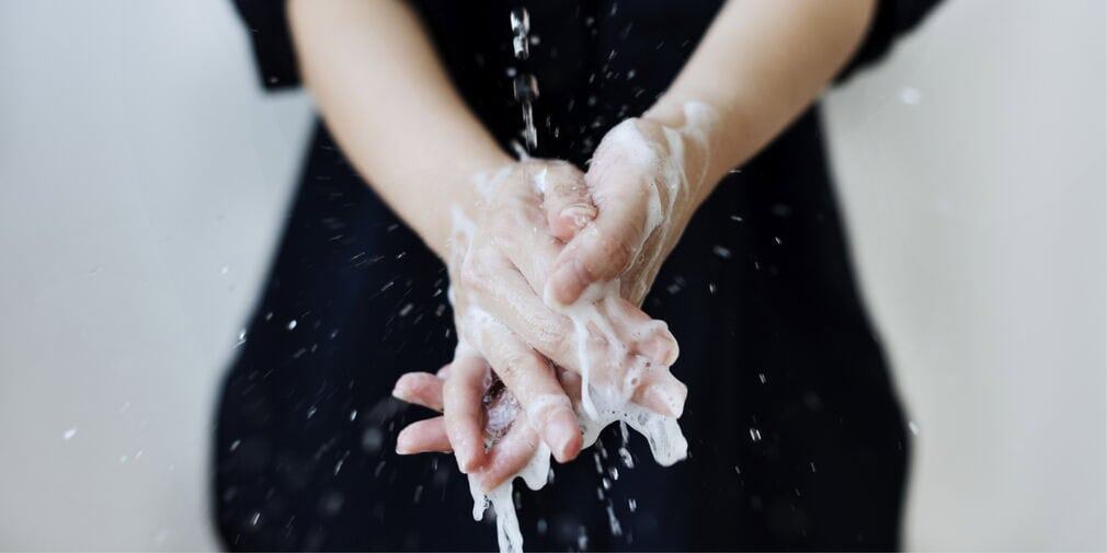 Soapy hands are in the foreground under a stream of water, while part of a woman in black is in the background. Restaurant hot water requirements need to be met to prevent scalding and burns.