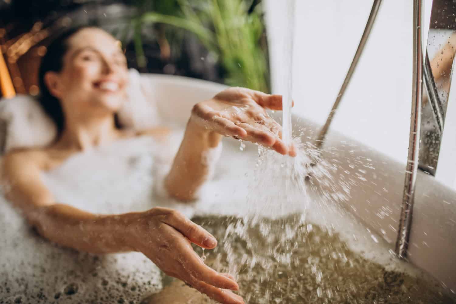 A woman enjoys a relaxing bath in a hotel bathtub, her hand splashes water from the running bathtub faucet. How to reduce hot water usage in hotels includes restroom water audits. 