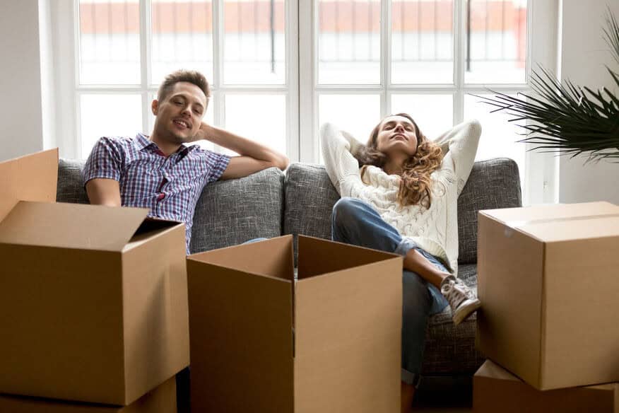 A young couple relax on a couch surrounded by empty moving boxes unpacked in their furnished apartment, with a large bright window behind them.