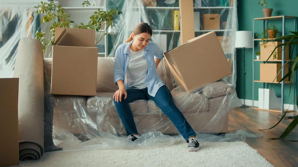 A woman sits on a plastic covered couch holding her back in pain after moving boxes and furniture into her new apartment.