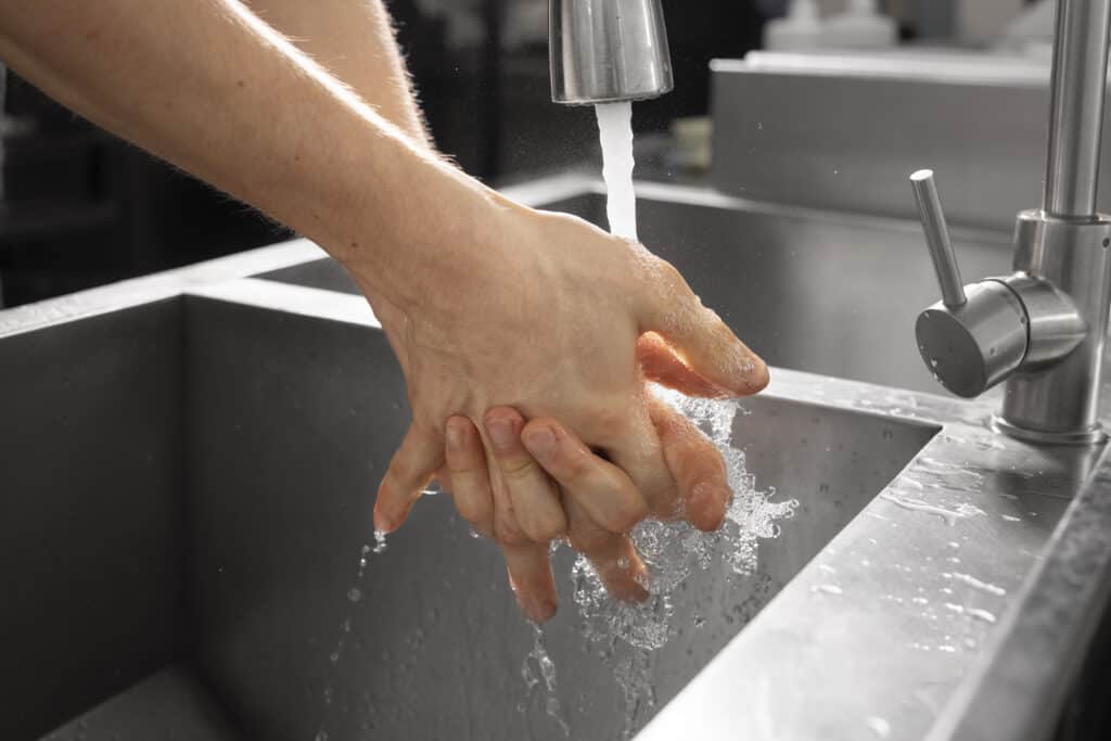A close up of hands washing under a running water faucet in a restaurants kitchen sink.