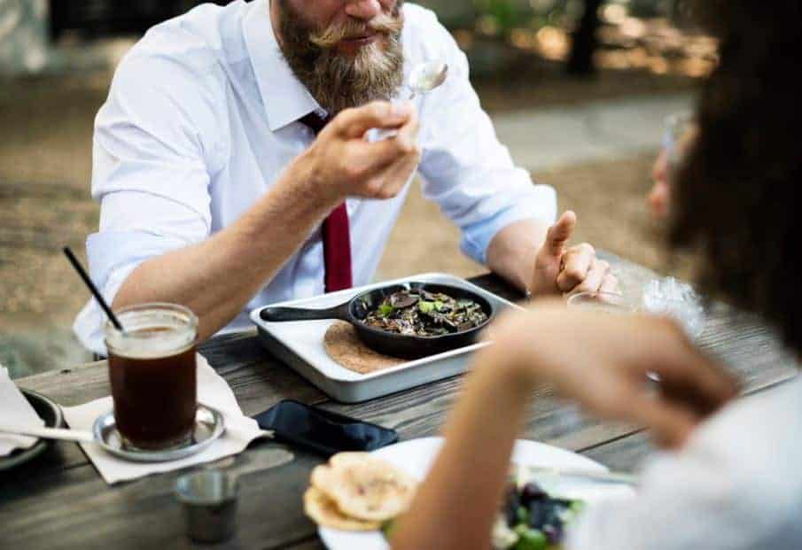 A man and woman sit outdoors, eating food and drinks at a wood table and talking.