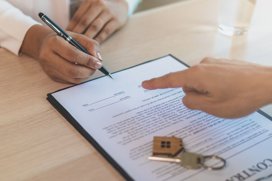 A landlord points to the part of the lease contract as the tenant gets ready to sign. A housekey attached to a wooden home keychain. 