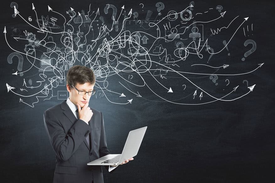 A man in a business suit rubs his chin while he looks at his laptop. The black wall behind him has chalk arrows, question marks and other symbols written around his head to imply brainstorming and thinking. 