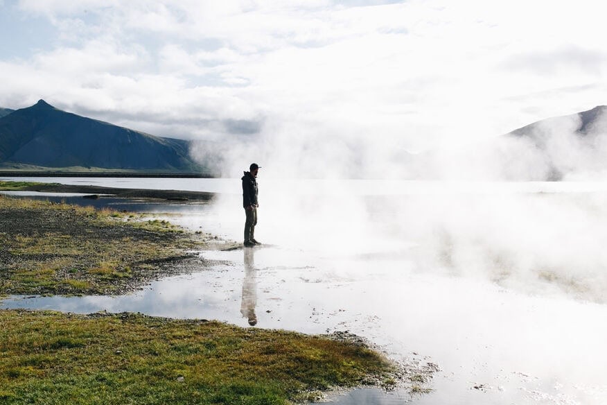 A man stands at the edge of a steaming geyser in Iceland, watching the hot water steam. 