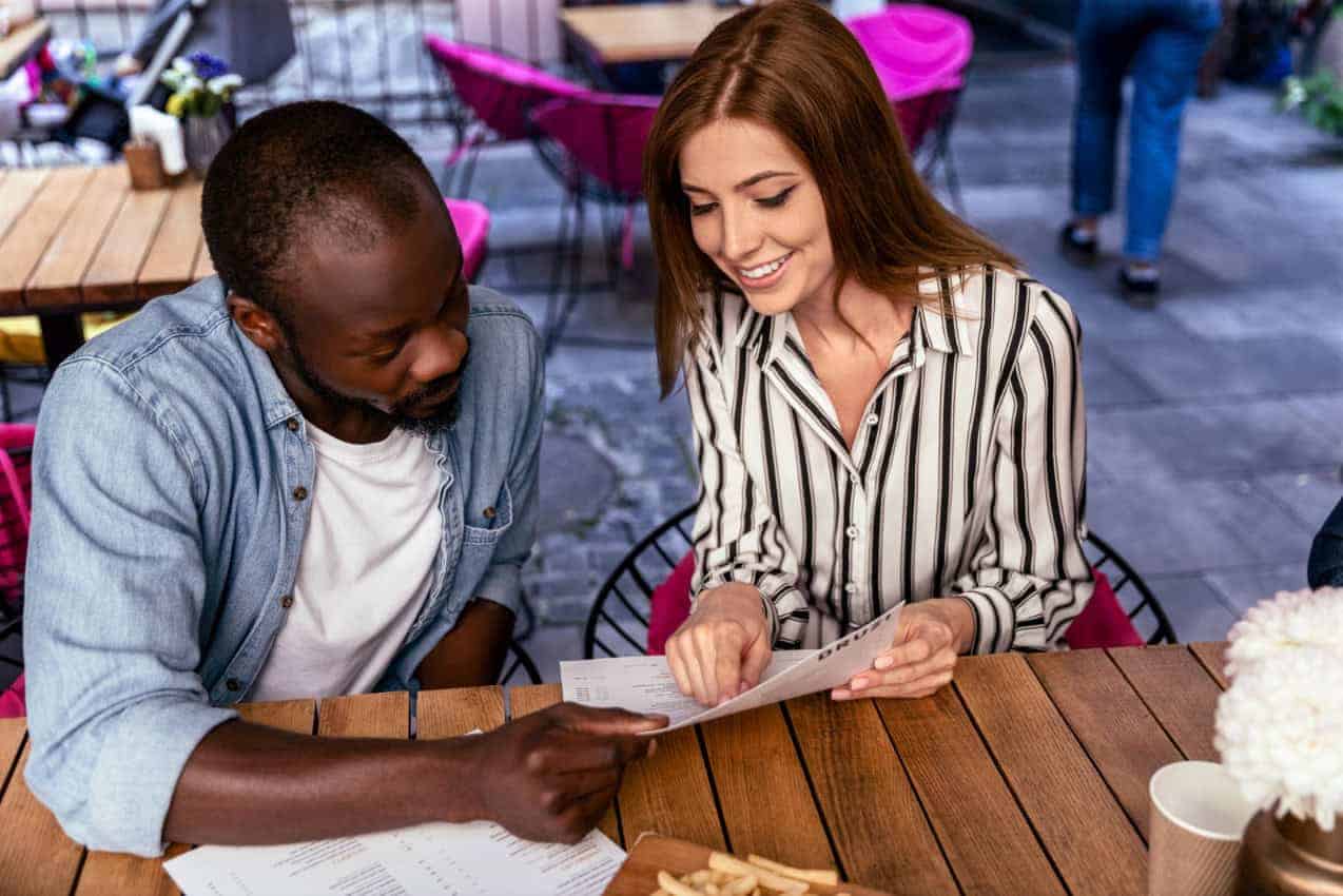 A couple reads a paper menu together while sitting on an outdoor restaurant patio.