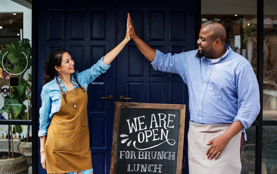 A man and woman stand outside a restaurant and high-five over a board that reads "We are open for brunch and lunch."