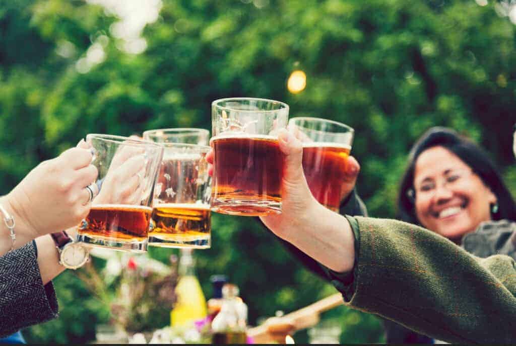 A close-up of pint glasses filled with beer clinking together in a toast outside in a beer garden. A woman participating in the cheers smiles.