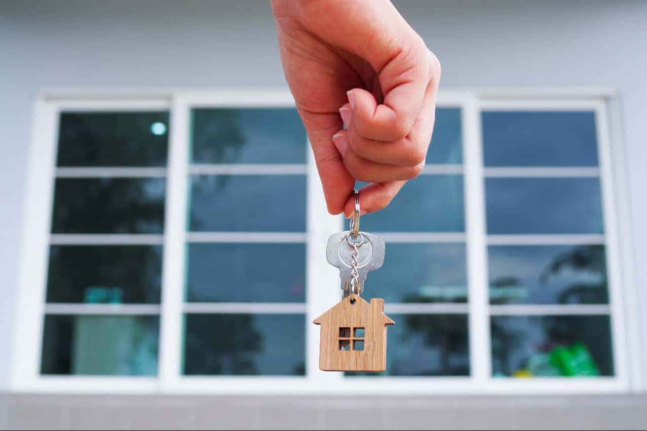 In front of large house windows, a manicured hand dangles keys with a small house token on the keyring.