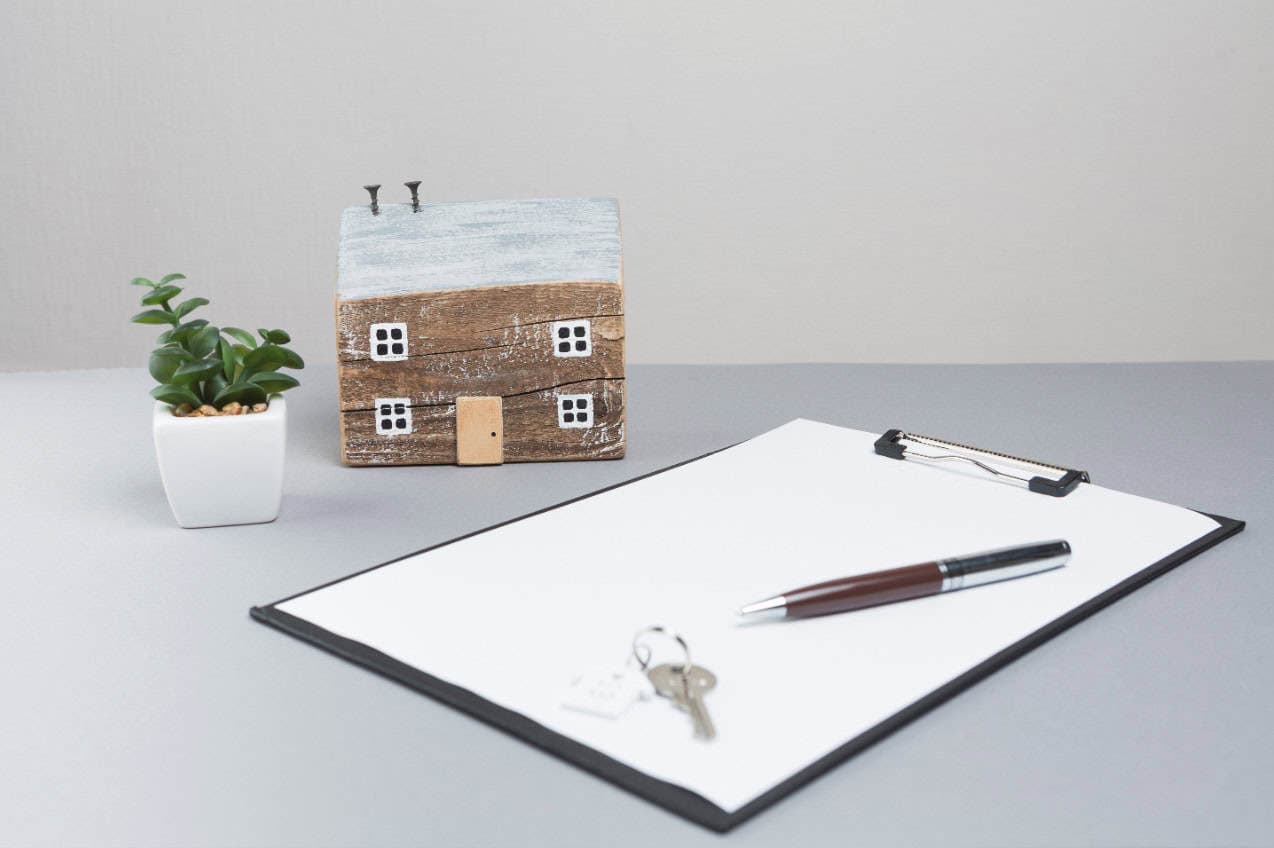 A miniature model of a home sits on a gray desk next to a small potted plant, a clipboard with a pen, and the house keys.