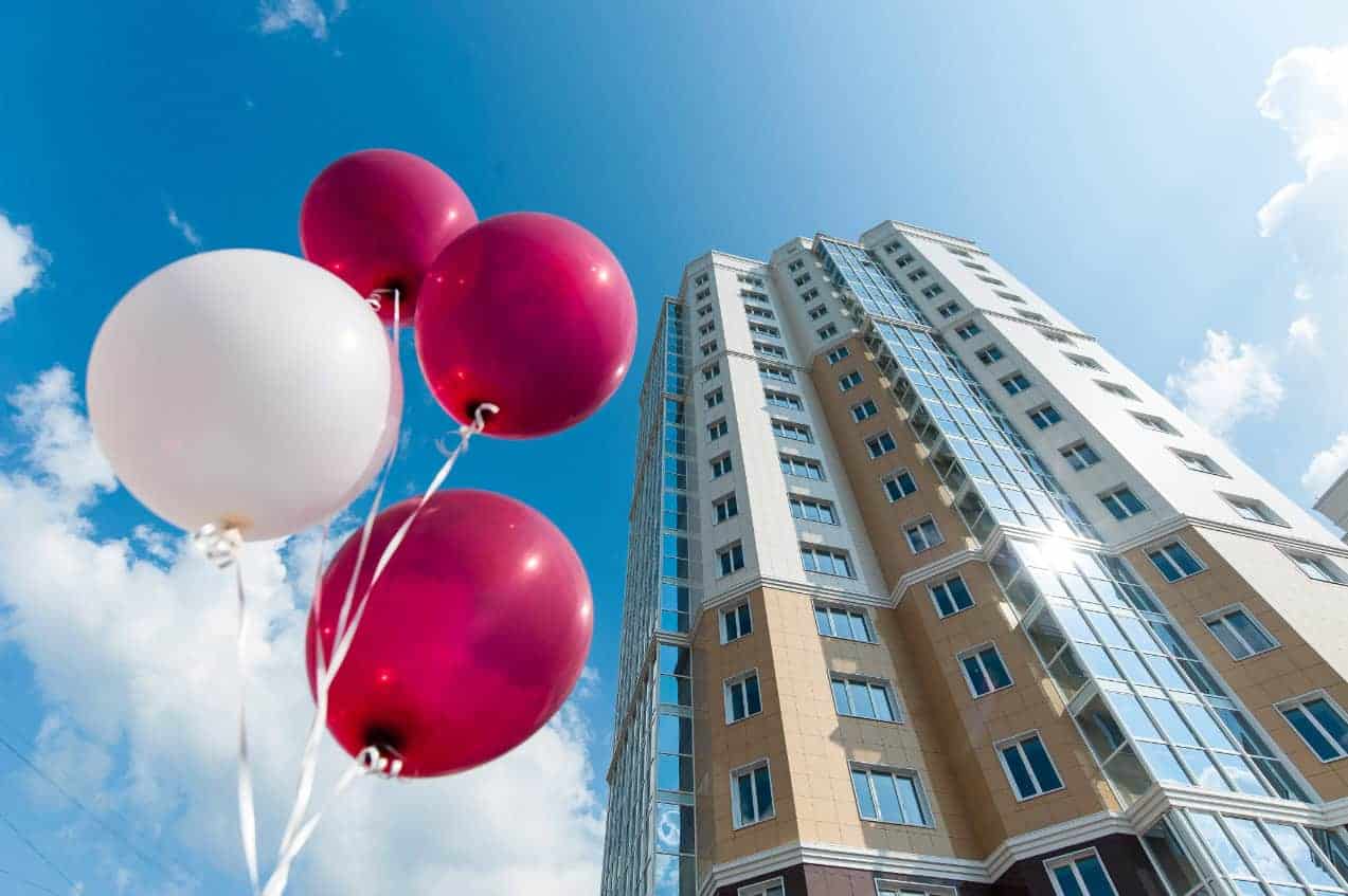 A photo of a tall apartment building stretching into the blue sky. Red and white balloons floating nearby allude to a new lease celebration.