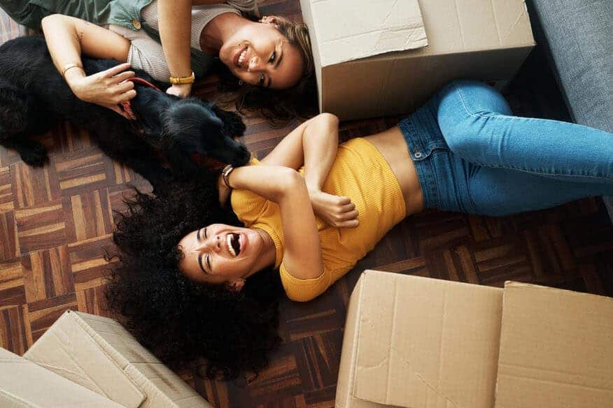 Two women and a dog lay on the wooden floor un their new apartment, surrounded by moving boxes. 