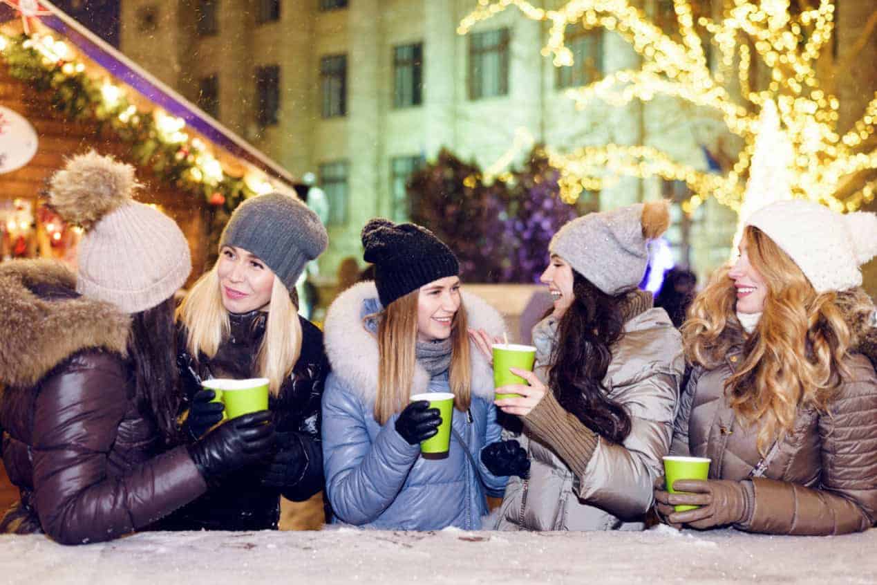 Five women dressed in winter gear sit at an outside table while drinking and having fun. 