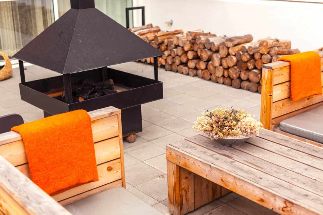 An outdoor dining space has a small, covered metal firepit next to wood furniture in the center. A wood pile is along the back wall, making this space cozy during winter. 