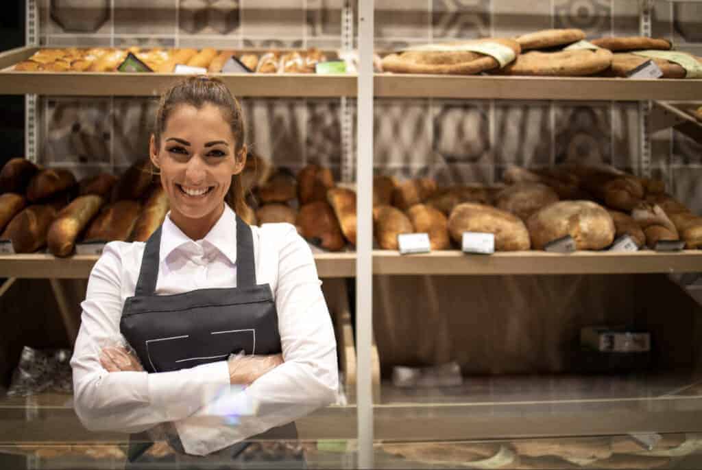 A smiling baker in a black apron stands proudly with her arms crossed in front of shelves full of loaves of bread.