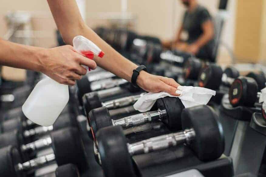 A female gym employee uses a spray bottle and a cloth to carry out gym cleaning best practices. 