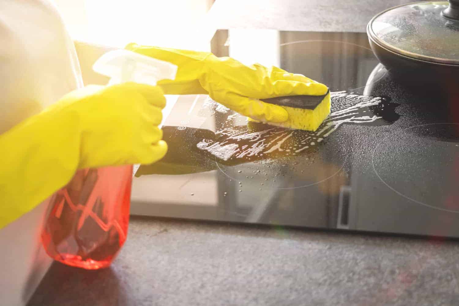 A cleaner's hands in bright yellow gloves are using a sponge and a spray bottle to clean a stovetop. Keep accessible cleaning supplies on hand to ensure all tasks on your restaurant cleaning checklist are completed.