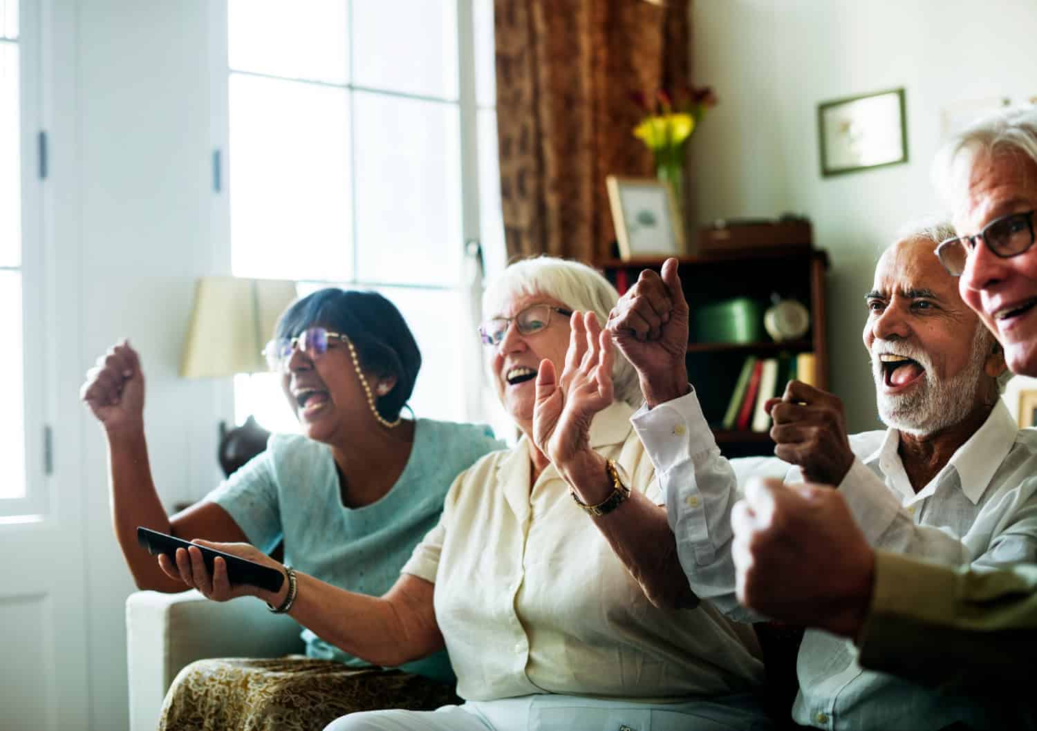 A group of seniors - two women and two men - sit on a couch, cheering. They hold their hands up in the air in celebration, all smiling.