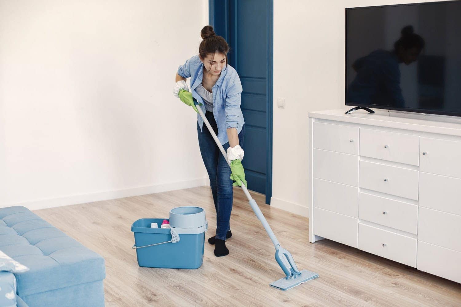 A young woman mops the floor in her apartment living room, making sure to clean up all signs of normal wear and tear on her apartment before moving out.