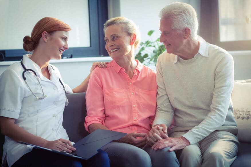 A smiling senior couple sit on a couch with their health care provider at an assisted living facility. 