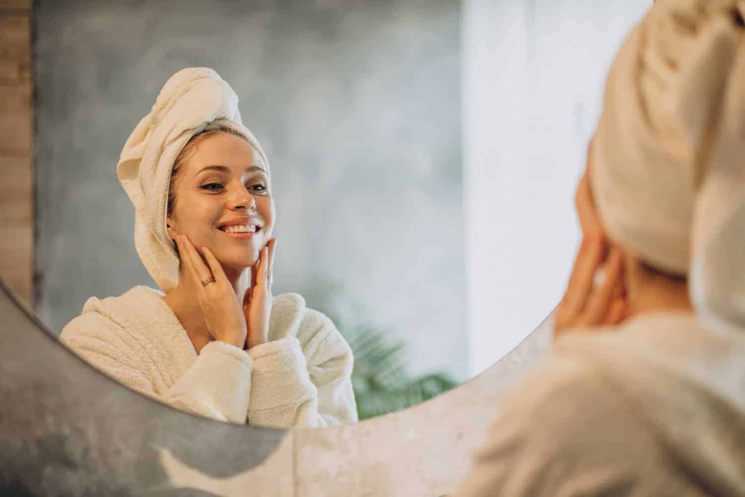 A woman in a robe with her hair wrapped in a towel smiles into the mirror as she caresses her face after a spa treatment.