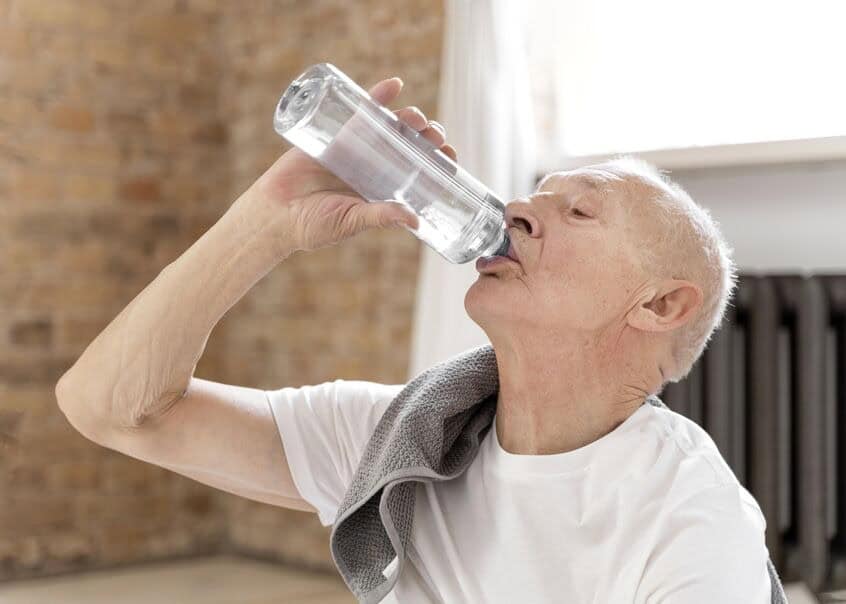 An elderly man tips his head back to drink from a bottle of water after a workout. 