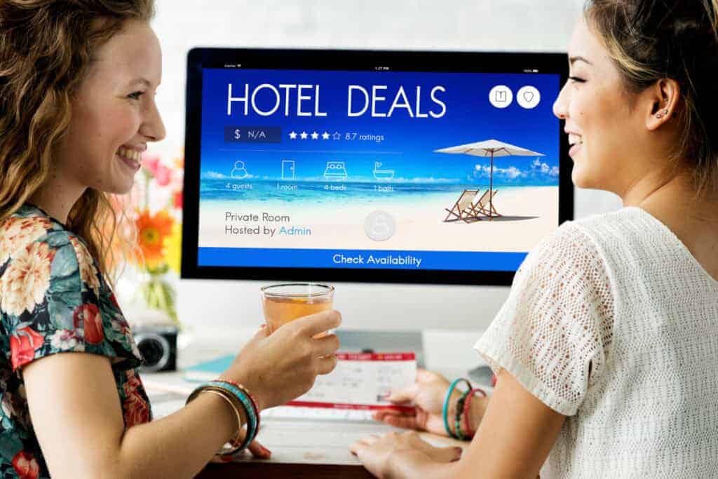 Two women smiling at each other while searching online for affordable hotel deals on a computer.